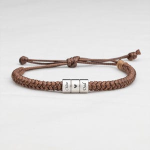 May include: A brown braided cord bracelet featuring three silver-toned cylindrical beads. The center bead displays a heart symbol, while the two flanking beads are engraved with the names Chloe and Dad, respectively, in a dark script font.