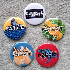 May include: Five round button badges with the word "PARXIE" printed in black on a white, yellow, blue, red, and green background. The badges feature a variety of designs, including a double dare logo, a frog, a fruit, and a group of people.
