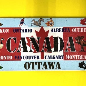 May include: Rectangular souvenir featuring a red and white Canadian flag design with the word "CANADA" in bold letters. The design includes illustrations of a goose, moose, and hockey player, with city and province names.