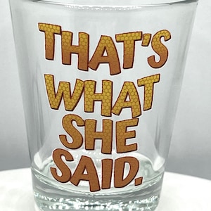 Peut inclure: Verre à shot transparent avec l'inscription "THAT'S WHAT SHE SAID" imprimée en lettres marron et jaune. Le verre est cylindrique avec une base épaisse et un bord légèrement évasé. Le texte présente un motif en nid d'abeille.