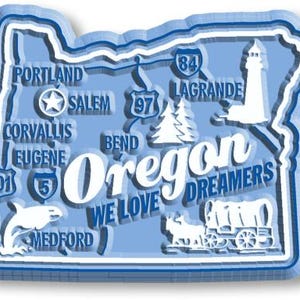 May include: A light blue, die-cut magnet in the shape of the state of Oregon. The magnet features white illustrations of a lighthouse, a whale, and a covered wagon. The word "Oregon" is prominently displayed, along with the phrase "We Love Dreamers."