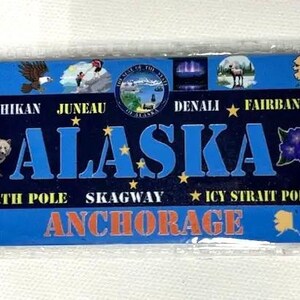 May include: A rectangular Alaska souvenir magnet with a blue background. The word "ALASKA" is in large blue letters. The magnet features illustrations of a bear, eagle, and other Alaskan imagery, along with city names like Anchorage.