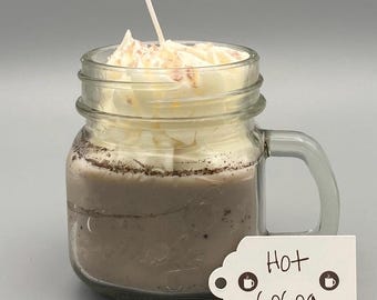 Puede incluir: Una vela de vidrio en forma de taza de chocolate caliente con crema batida y una mecha blanca. El tarro tiene una manija y una etiqueta blanca que dice "Chocolate caliente" con un icono de taza de café a cada lado.