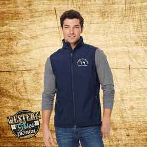 May include: A navy blue zip-up vest featuring a white embroidered logo on the chest that reads Larsen Ranches FY Colorado. The vest is worn over a grey long-sleeve shirt and blue jeans, showcasing a high collar and two side zippered pockets.