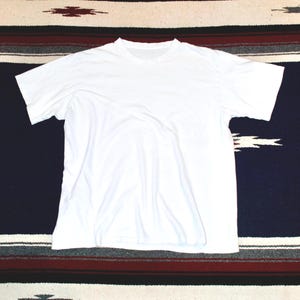May include: A plain white, short-sleeved t-shirt. The shirt is made of a soft material and has a crew neck. The shirt is laying flat on a patterned textile with navy blue, red, and white stripes.