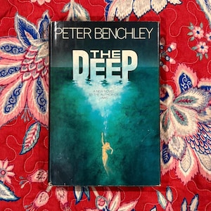 May include: A hardcover book titled "The Deep" by Peter Benchley. The cover features a teal and blue underwater scene with a diver and the title in white. The book is on a red floral patterned background.