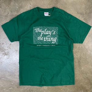 以下が含まれることがあります： 白文字で「The play's the thing」とシェイクスピアの戯曲をいくつかリストした緑色のTシャツ。テキストには「Georgia Shakespeare Festival」も含まれています。