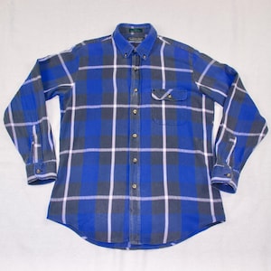 Puede incluir: Una camisa de franela a cuadros azul, gris y blanco con cuello abotonado. La camisa de manga larga tiene un bolsillo en el pecho y puños abotonados. La camisa está hecha de una tela suave y tejida.