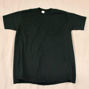 Könnte beinhalten: Ein dunkelgrünes T-Shirt mit kurzen Ärmeln und einer Brusttasche. Das Shirt besteht aus einem weichen Material und hat einen Rundhalsausschnitt. Das Kleidungsstück liegt flach auf einem neutralen Hintergrund.