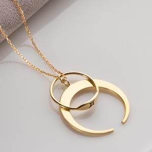 May include: A gold-colored necklace featuring a delicate chain and a pendant with two interlocking shapes: a circle and a crescent moon. The moon shape has a small symbol engraved on it. The necklace is displayed on a white surface.