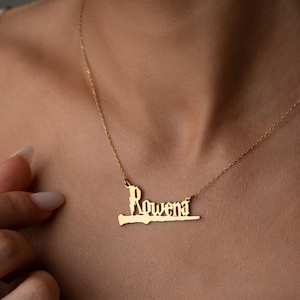 Könnte beinhalten: Eine goldfarbene Halskette mit dem Namen "Rowena" in einer stilisierten Schriftart. Der Anhänger hängt an einer zarten Kette. Die Halskette wird am Hals einer Person gezeigt.