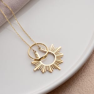 May include: A gold-colored necklace with a sunburst pendant and a delicate chain. The pendant features a circular design with radiating lines, and a smaller circle is attached. The necklace is displayed on a white surface.