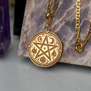 May include: A gold chain necklace with a round pendant featuring a pentacle with a five-pointed star, moon, and flames. The pendant is on a marble surface.
