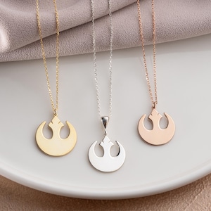 May include: Three necklaces with pendants in gold, silver, and rose gold. Each pendant has a circular design with a stylized emblem, hanging from a fine chain. The necklaces are displayed on a white surface.