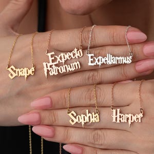 May include: A collection of gold and silver nameplate necklaces. The necklaces feature names and phrases like "Snape", "Expecto Patronum", and "Expelliarmus" in a stylized font. The necklaces are displayed on a person's hand.
