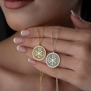 以下が含まれることがあります： 金色のペンダントネックレスと銀色のペンダントネックレスの2つ。それぞれ円形のフレームの中にカイ・ローのシンボルが描かれています。ペンダントは繊細なチェーンから吊り下げられています。ぼやけた背景にネックレスが配置されています。