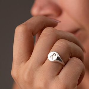 以下が含まれることがあります： 丸い面に文字「R」が刻まれたシルバーのシグネットリング。指にはめています。背景はぼやけており、リングと手に焦点が当てられています。