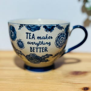 Könnte beinhalten: Cremefarbene Keramik-Teetasse mit blauen Blumenmustern und dem Text "TEA makes everything BETTER". Der Henkel und der Boden sind blau. Die Tasse steht auf einer Holzoberfläche.
