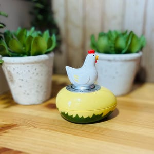 May include: A decorative kitchen timer shaped like an egg, with a white chicken on top. The egg-shaped timer is yellow with a green base. The timer has a silver dial and is set against a blurred background with potted plants.