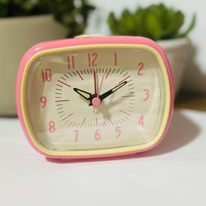 May include: A rectangular analogue alarm clock with a pink outer frame and a pale yellow inner border. The clock face is off-white with pink Arabic numerals from 1 to 12. It features black hour and minute hands with luminous tips and a thin red second hand.