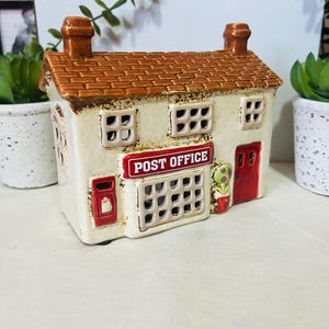May include: A ceramic post office ornament with a cream exterior, brown tiled roof, and red details. The building has a sign reading "POST OFFICE", a red door, and small windows. A decorative item for home decor.