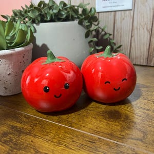 Peut inclure: Deux salières et poivrières en céramique rouge en forme de tomate, avec des tiges vertes et des visages souriants. Les salières et poivrières sont posées sur une surface en bois, avec une plante et une enseigne "Rosewood" en arrière-plan.
