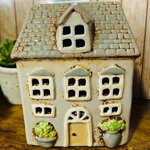 May include: A ceramic house-shaped candle holder with a light blue facade, a gray tiled roof, and multiple windows. The house has a small dormer window and a yellow door. Two small green potted plants are on the front. The sign says "Rosewood".