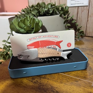 May include: A fish-shaped corkscrew with a wooden handle and stainless steel accents, presented in a blue box. The box contains a card with the text "SPIRIT OF ADVENTURE" and a red fish illustration. A decorative item for wine enthusiasts.