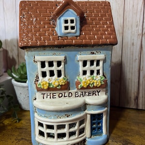Könnte beinhalten: Eine Keramikminiatur einer Bäckerei mit braunem Dach, blauen Wänden und weißen Fensterrahmen. Der Text "THE OLD BAKERY" ist über dem vorderen Fenster zu sehen. Gelbe Blumen befinden sich in den Blumenkästen. Das Gebäude hat eine kleine blaue Tür.