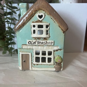 Puede incluir: Un portavelas de cerámica con forma de casa de color turquesa con tejado y chimenea marrones. La casa presenta una ventana con marco blanco, una puerta y el texto "OLD TEASHOP". Una pequeña planta en maceta está en el lateral.