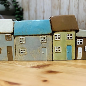 May include: A ceramic house planter featuring a row of colorful houses. Each house has a different colored roof, including white, blue, and brown. The houses have square windows and doors, and are set on a wooden surface.