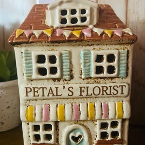 May include: A ceramic house-shaped container with the text "PETAL'S FLORIST". The house has a brown roof, white walls, and colorful accents. The windows have blue shutters, and the door has a heart-shaped cutout.