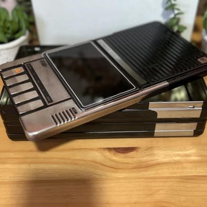 May include: A black and silver metal box with a hinged lid. The lid has a black, ridged surface and a silver panel with buttons and a screen. The box is sitting on top of a stack of similar black boxes. The boxes are on a wooden surface.