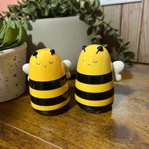 Può includere: Due saliere e pepiere in ceramica a forma di ape. Le saliere sono gialle con strisce nere, piccole ali bianche e facce sorridenti. Lo sfondo comprende vegetazione e una superficie in legno.
