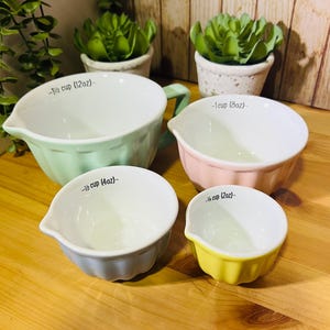 Op de afbeelding: Een set van vier pastelkleurige keramische maatbekers. De bekers zijn in tinten mintgroen, roze, grijs en geel. Elke beker heeft een witte binnenkant en een schenktuit. De maten staan in zwarte tekst aangegeven.