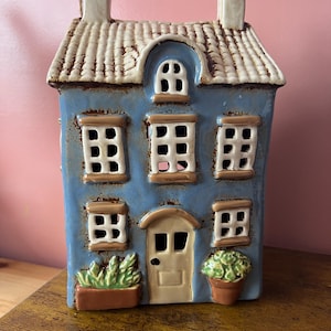 Puede incluir: Un portavelas de cerámica azul con forma de casa con tejado texturizado y chimeneas. La casa tiene varias ventanas, una puerta principal y plantas en macetas decorativas. El diseño general es rústico y encantador.