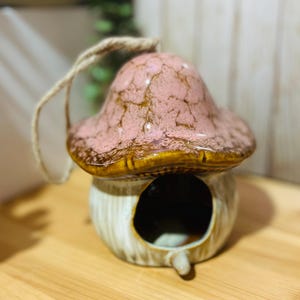 May include: A ceramic mushroom-shaped birdhouse with a pink and brown cap and a cream-coloured stem. The birdhouse has a round opening and a jute rope for hanging. The design is whimsical and decorative.