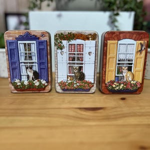 May include: Three decorative metal tins featuring cat scenes. Each tin depicts a window with a cat, shutters, and a flower box. The tins have different color schemes: blue, white, and yellow. The tins are on a wooden surface.