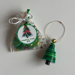 May include: A festive Christmas tree kit with a green and gold color scheme. The kit includes a clear bag with gold stars, a tag with a colorful tree design, and green buttons. A separate tree ornament made of green buttons and a gold ring is also present.