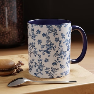 May include: A white ceramic mug with a blue floral pattern and a dark blue rim and handle. The mug is sitting on a wooden surface.