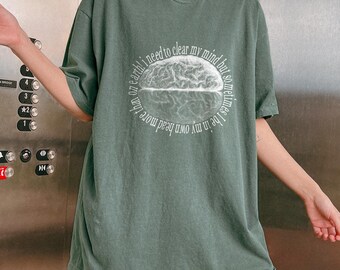 May include: Sage green t-shirt with a circular graphic of a brain and the text "I need to clear my mind but sometimes I'd be in my own head more than on earth!" The shirt has short sleeves and a relaxed fit.