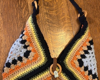 May include: A handmade crochet bag with a colorful granny square design. The bag features a navy blue strap and wooden rings. The color palette includes shades of blue, orange, yellow, and black. A small tassel and wooden ring detail the front.