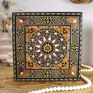 Può includere: Scatola quadrata in legno con un intricato design a intarsio geometrico. La scatola presenta un motivo centrale a stella con elementi bianchi, marroni, gialli e neri. Il bordo presenta forme geometriche ripetute e una cornice scura.