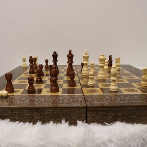 Puede incluir: Un juego de ajedrez de madera con detalles tallados. El tablero es marrón y beige, con piezas de ajedrez en tonos de madera claros y oscuros. Algunas piezas están caídas, lo que sugiere un juego en curso.