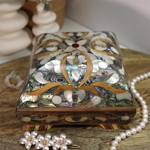 Op de afbeelding: Een rechthoekige sieradendoos met een decoratief bloemmotief. De doos is gemaakt van hout en ingelegd met iriserende abalone schelp en parelmoer. De doos heeft een scharnierend deksel en staat op kleine voetjes. Een parelketting en bloemen haarclip staan op de voorgrond.