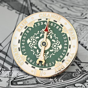 May include: A round, decorative weather gauge pin with a green center and gold accents. The pin features a needle pointing to "FAIR" and the words "STORMY", "RAIN", "CHANGE", and "VERY DRY". Numbers and decorative patterns are also visible.