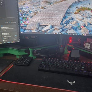 Peut inclure: Configuration informatique avec deux écrans, un clavier noir et un tapis de souris avec un bord rouge lumineux. Les écrans sont montés sur des supports verts. L'arrière-plan affiche un paysage.