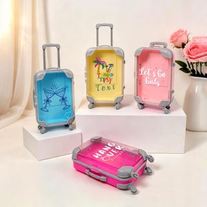 May include: Four miniature, colourful suitcases with clear sides and grey accents. One is blue with champagne glasses, one yellow with a palm tree graphic, one pink with "Let's Go Girls", and one pink with "Hang Over".