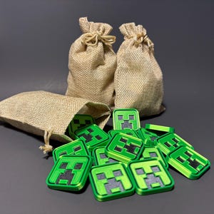 May include: Three small burlap bags with drawstrings, two are filled with green and black Minecraft Creeper face figurines. The bags are on a gray surface.