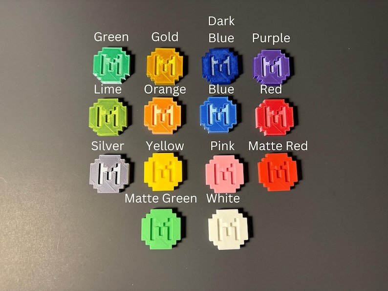 May include: A set of 14 round, plastic, 3D printed tokens in various colors, including green, gold, dark blue, purple, lime, orange, blue, red, silver, yellow, pink, matte red, matte green, and white. Each token features a stylized letter "M" in the center.